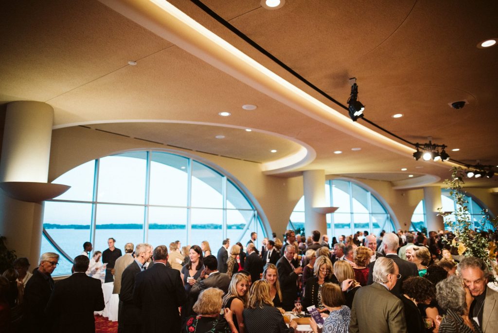 A large event space with high ceilings and large windows overlooking Lake Monona filled with a hundred people