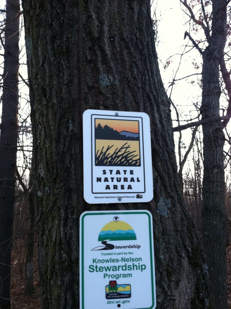 Two signs pinned to a tree that read "State Natural Area" and "Funded in part by the Knowles-Nelson Stewardship Program"